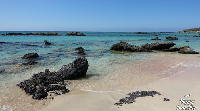 Το επικό οδοιπορικό μας στην Δυτική Κρήτη | Νομός Χανίων Η παραλία Ελαφονήσι στην Κρήτη με ροζ άμμο και κρυστάλλινα, ρηχά τιρκουάζ νερά, περιτριγυρισμένη από χαμηλούς λόφους κάτω από έναν ηλιόλουστο ουρανό.