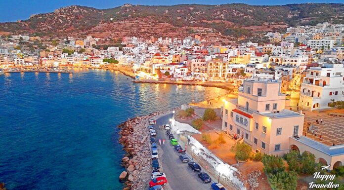 Παραθαλάσσια άποψη των Πηγαδιών Καρπάθου στο σούρουπο, με τα φώτα της πόλης να καθρεφτίζονται στη θάλασσα.