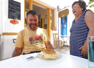 Ο Ευτύχης κάθεται στο τραπέζι του παραδοσιακού καφενείου "Κεντρικόν" στην Τήνο με ένα γαλακτομπούρεκο μπροστά του, ενώ στέκεται δίπλα του η κυρία που το έφτιαξε.