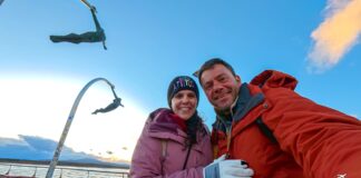 Ευτύχης Μπλέτσας και Ηλέκτρα Αστέρη στο Monumento al Viento στο Puerto Natales, ποζάρουν μπροστά από το εμβληματικό γλυπτό που συμβολίζει τους ισχυρούς ανέμους της Παταγονίας, με φόντο το παραλιακό μέτωπο και το φιόρδ Última Esperanza